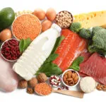 Vista dall’alto di fonti proteiche diverse su fondo bianco: petto di pollo, salmone, uova, legumi, formaggio, frutta secca, riso e broccoli