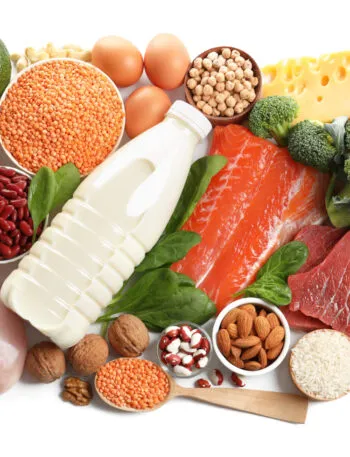 Vista dall’alto di fonti proteiche diverse su fondo bianco: petto di pollo, salmone, uova, legumi, formaggio, frutta secca, riso e broccoli