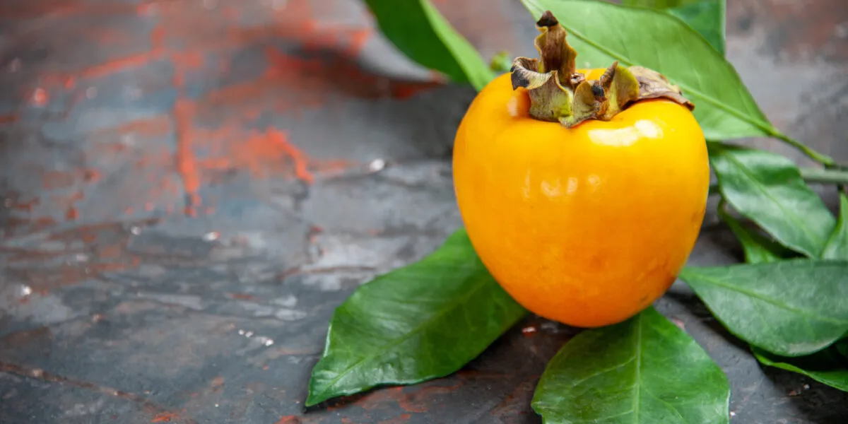 Un cachi arancione intero con il picciolo e alcune foglie verdi, appoggiato su un piano di ardesia scura