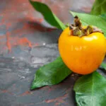 Un cachi arancione intero con il picciolo e alcune foglie verdi, appoggiato su un piano di ardesia scura