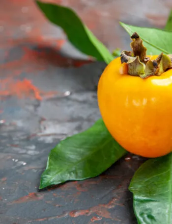 Un cachi arancione intero con il picciolo e alcune foglie verdi, appoggiato su un piano di ardesia scura