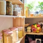 Dispensa in legno con barattoli di vetro contenenti riso, pasta, lenticchie e legumi secchi, accanto a un cesto di mele