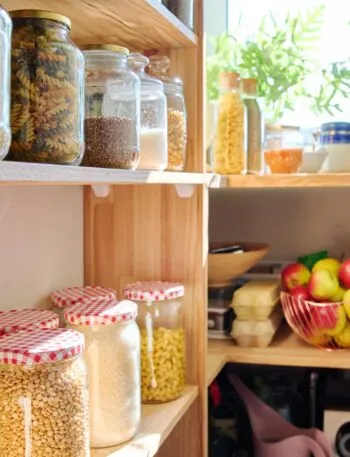 Dispensa in legno con barattoli di vetro contenenti riso, pasta, lenticchie e legumi secchi, accanto a un cesto di mele
