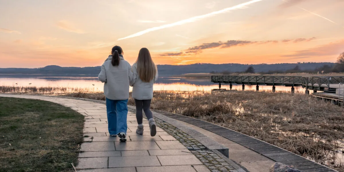 Due persone che camminano di spalle su un vialetto lungo l'acqua al tramonto, con abbigliamento comodo e passo tranquillo