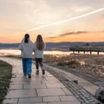 Due persone che camminano di spalle su un vialetto lungo l'acqua al tramonto, con abbigliamento comodo e passo tranquillo