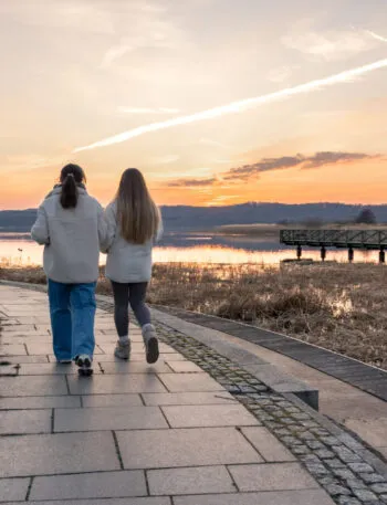 Due persone che camminano di spalle su un vialetto lungo l'acqua al tramonto, con abbigliamento comodo e passo tranquillo