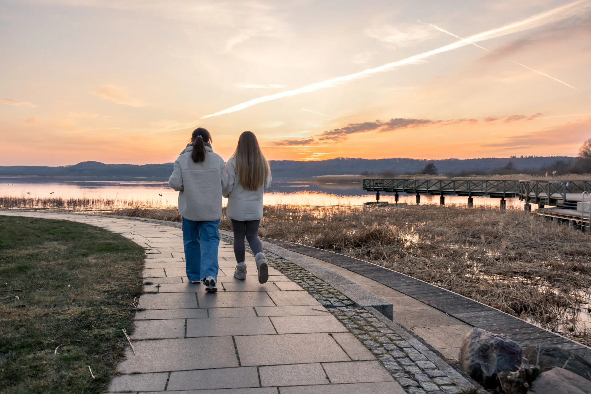 Due persone che camminano di spalle su un vialetto lungo l'acqua al tramonto, con abbigliamento comodo e passo tranquillo