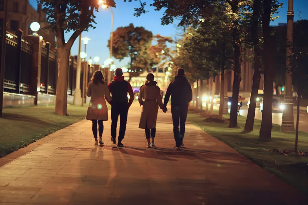 Quattro persone che camminano di spalle lungo un viale alberato illuminato dai lampioni, all'imbrunire