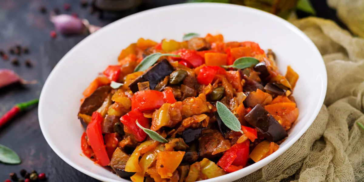 Caponata di melanzane a cubi grandi con peperoni rossi e gialli, pomodoro e capperi, servita in un piatto bianco fondo