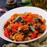Caponata di melanzane a cubi grandi con peperoni rossi e gialli, pomodoro e capperi, servita in un piatto bianco fondo