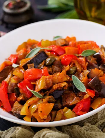 Caponata di melanzane a cubi grandi con peperoni rossi e gialli, pomodoro e capperi, servita in un piatto bianco fondo