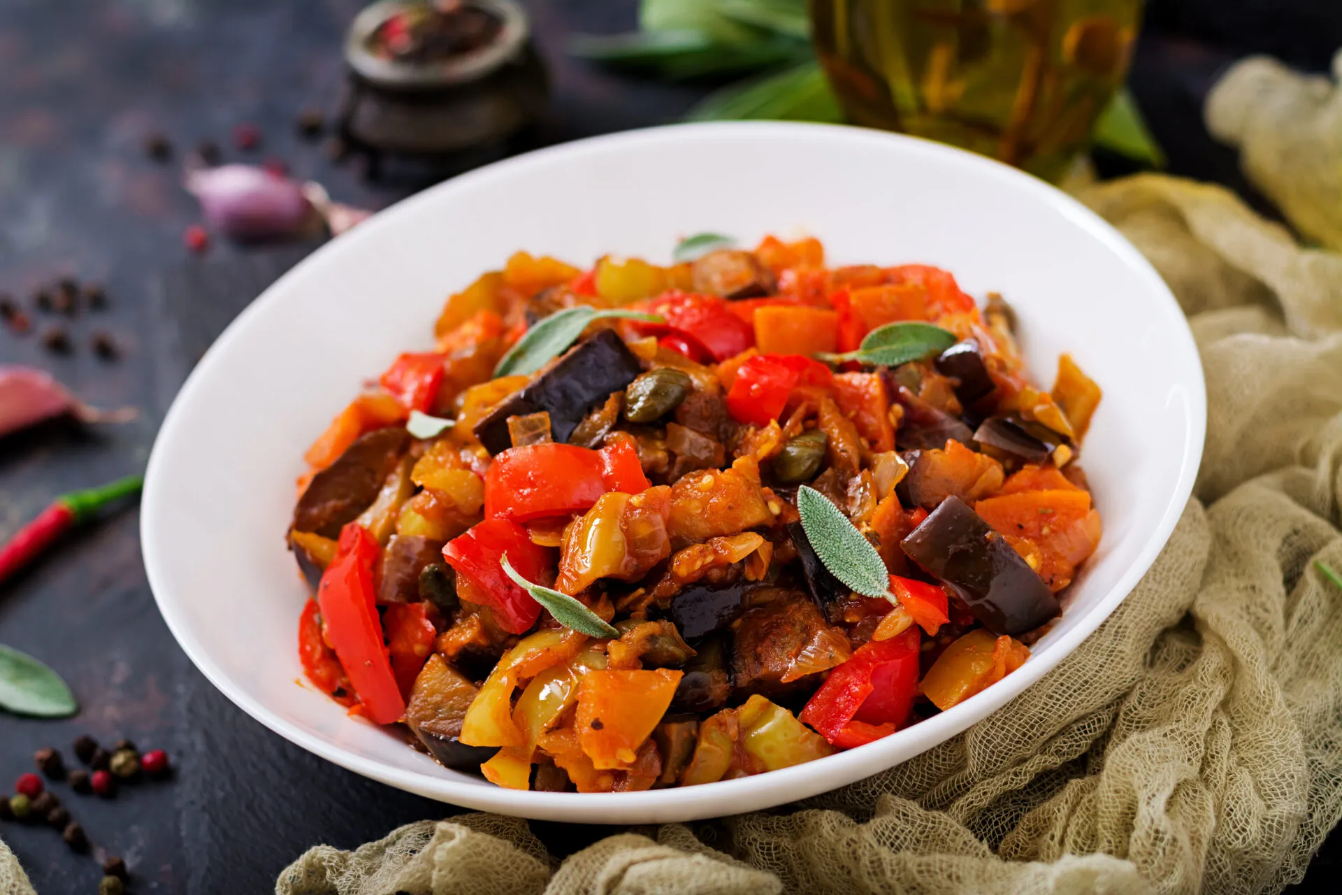 Caponata di melanzane a cubi grandi con peperoni rossi e gialli, pomodoro e capperi, servita in un piatto bianco fondo
