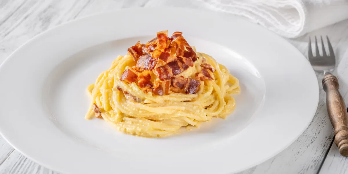 Piatto di spaghetti alla carbonara con crema liscia e lucida che avvolge la pasta, con guanciale croccante sopra