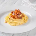 Piatto di spaghetti alla carbonara con crema liscia e lucida che avvolge la pasta, con guanciale croccante sopra
