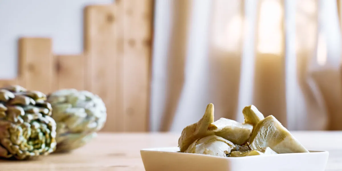 Piattino bianco con spicchi di carciofo cotti, con due carciofi interi appoggiati sul tavolo di legno chiaro sullo sfondo