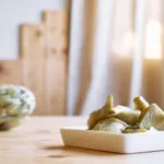 Piattino bianco con spicchi di carciofo cotti, con due carciofi interi appoggiati sul tavolo di legno chiaro sullo sfondo