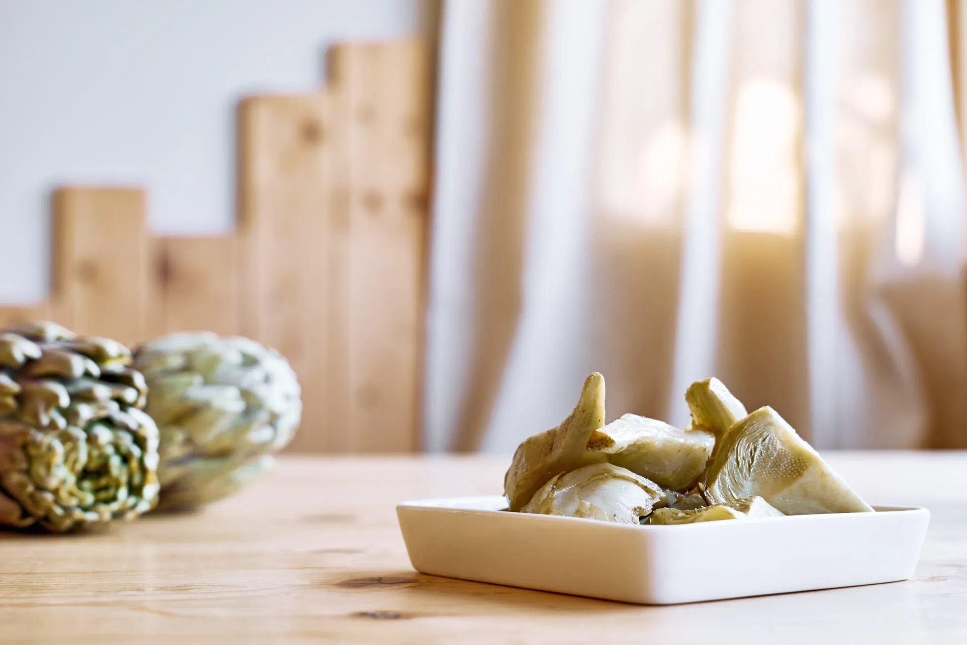 Piattino bianco con spicchi di carciofo cotti, con due carciofi interi appoggiati sul tavolo di legno chiaro sullo sfondo