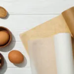Due rotoli di carta forno, uno bianco e uno avana, con fogli già tagliati accanto a uova, zucchero di canna e cucchiai di legno su un tavolo bianco