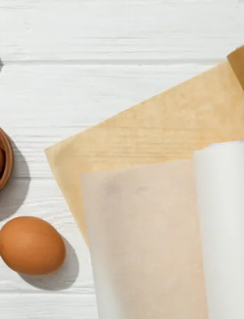 Due rotoli di carta forno, uno bianco e uno avana, con fogli già tagliati accanto a uova, zucchero di canna e cucchiai di legno su un tavolo bianco