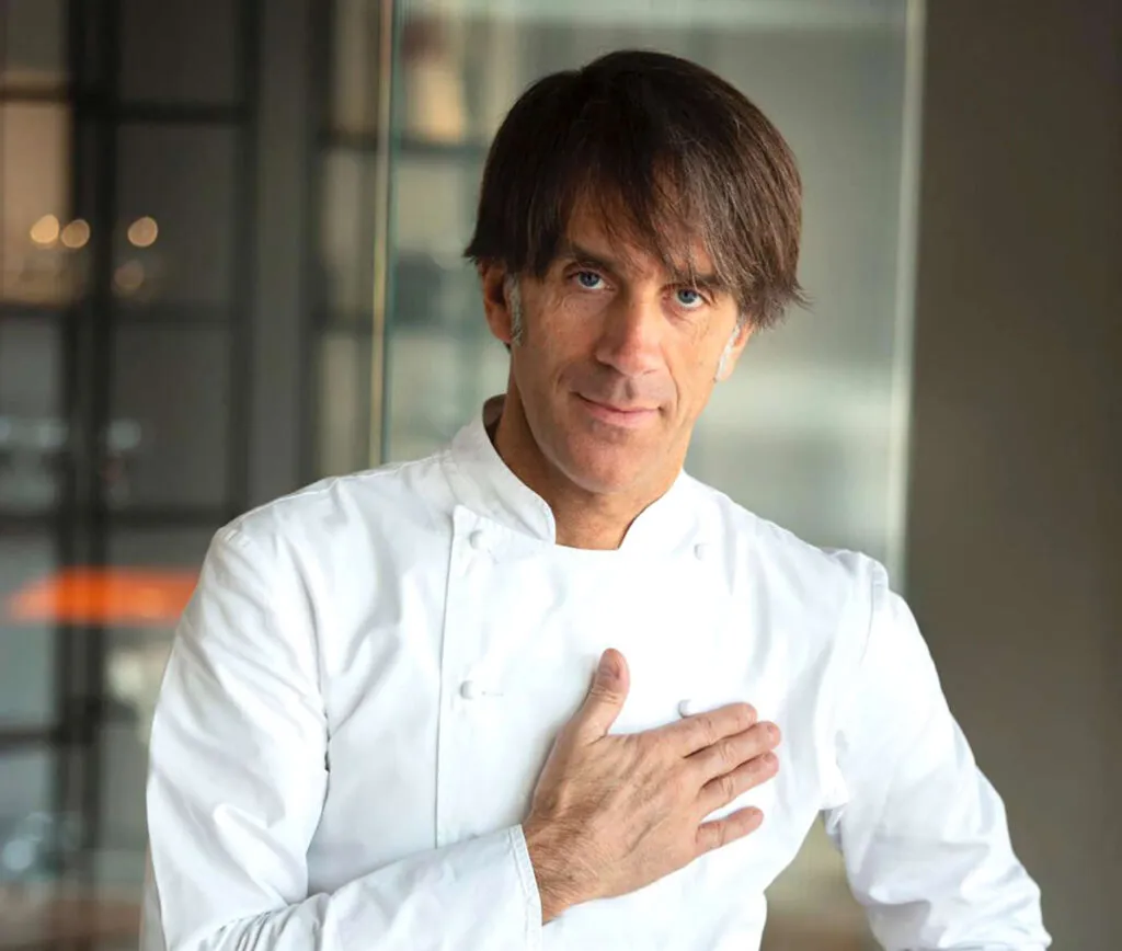 Ritratto dello chef Davide Oldani in giacca bianca da cucina, con la mano appoggiata sul petto, in un ambiente dalle linee moderne