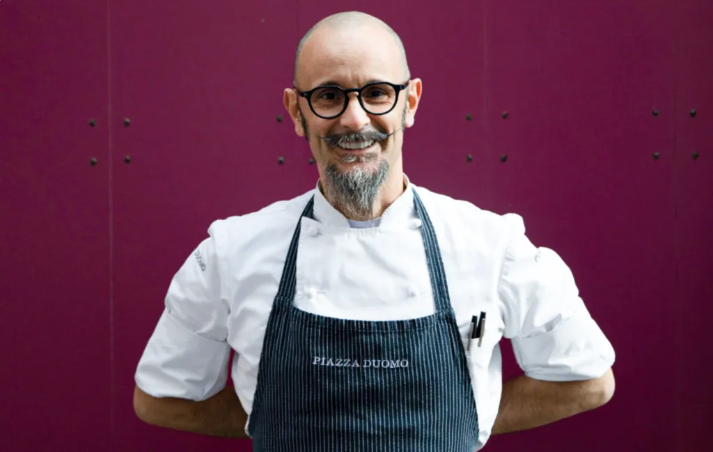 Lo chef Enrico Crippa in giacca bianca e grembiule a righe con il nome del ristorante, su fondo bordeaux