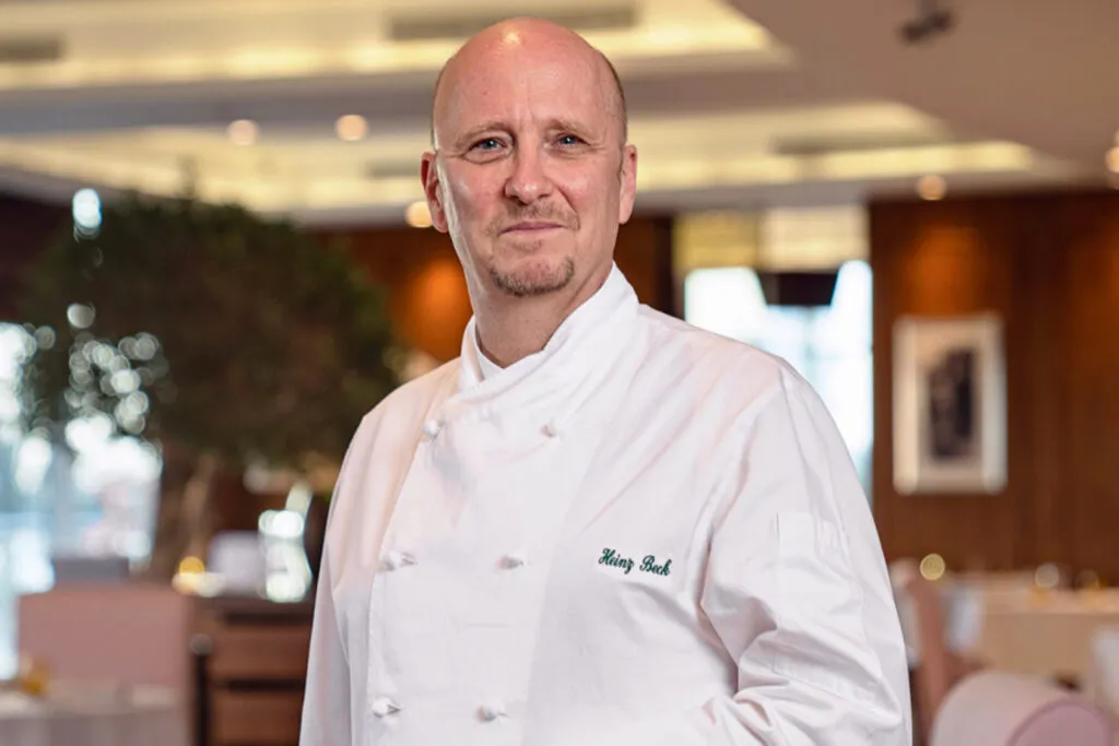 Lo chef Heinz Beck in giacca bianca con il nome ricamato, in piedi nella sala di un ristorante