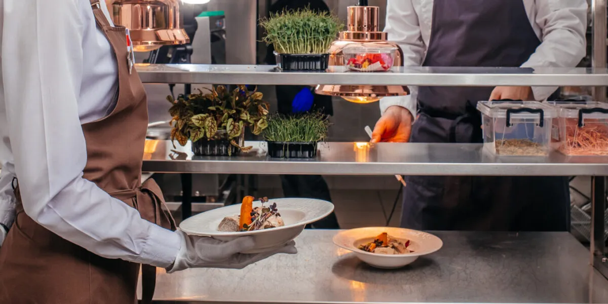 Cuoca in grembiule che porta un piatto impiattato al pass di una cucina professionale, con vaschette di germogli e micro-erbe sul ripiano