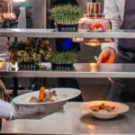 Cuoca in grembiule che porta un piatto impiattato al pass di una cucina professionale, con vaschette di germogli e micro-erbe sul ripiano