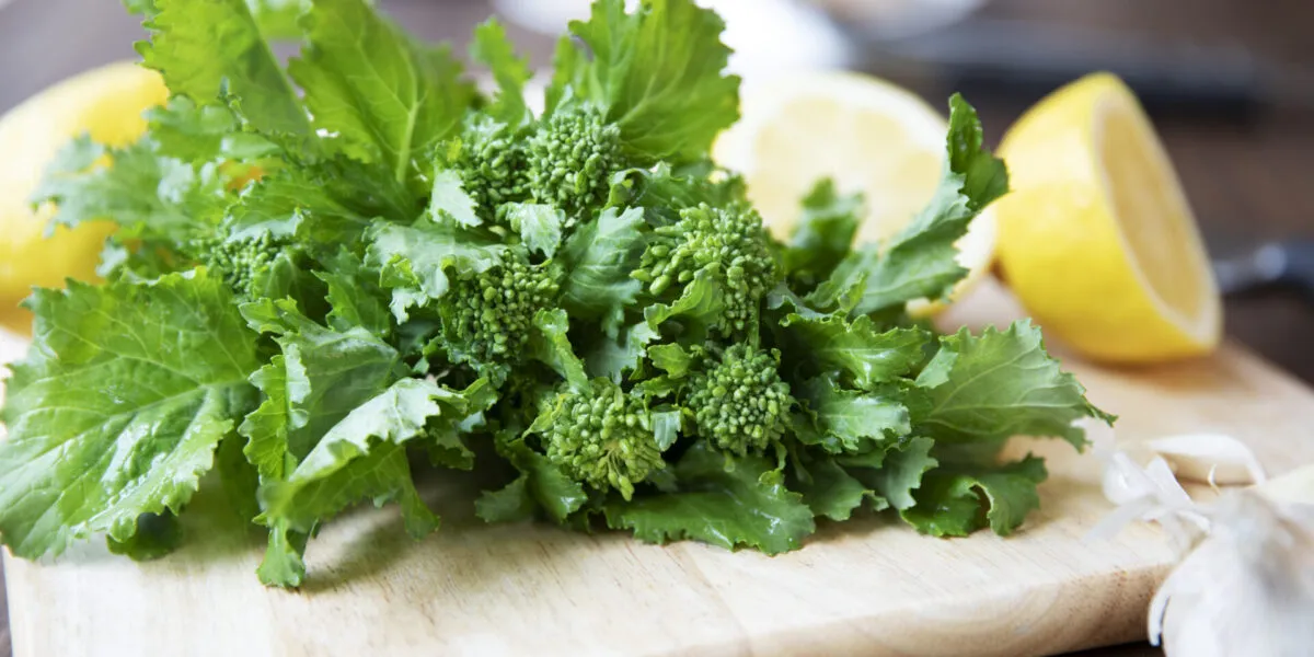 Mazzo di cime di rapa fresche con foglie intere e cimette su un tagliere di legno, accanto ad aglio e limone