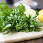 Mazzo di cime di rapa fresche con foglie intere e cimette su un tagliere di legno, accanto ad aglio e limone