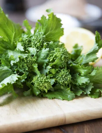 Mazzo di cime di rapa fresche con foglie intere e cimette su un tagliere di legno, accanto ad aglio e limone