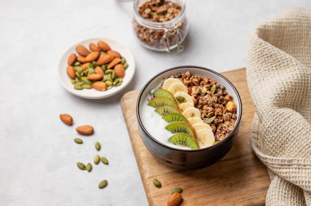 Ciotola di yogurt greco con kiwi, banana, granola e semi, accanto a un piattino di mandorle e semi di zucca