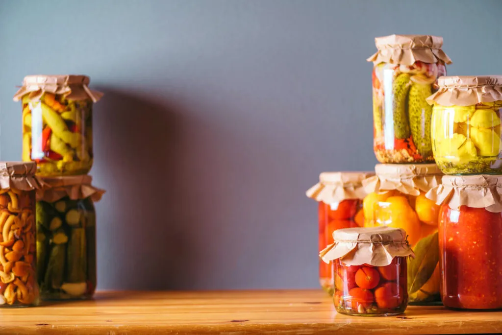 Vasetti di conserve fatte in casa con verdure sott'olio e sottaceto, chiusi con carta e appoggiati su un tavolo di legno