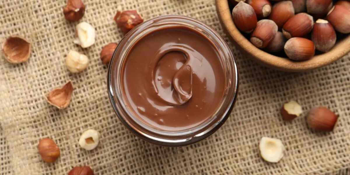 Barattolo di vetro con crema spalmabile alle nocciole vista dall'alto, circondato da nocciole intere e sgusciate su un telo di juta