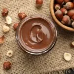 Barattolo di vetro con crema spalmabile alle nocciole vista dall'alto, circondato da nocciole intere e sgusciate su un telo di juta