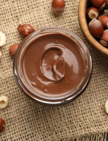 Barattolo di vetro con crema spalmabile alle nocciole vista dall'alto, circondato da nocciole intere e sgusciate su un telo di juta