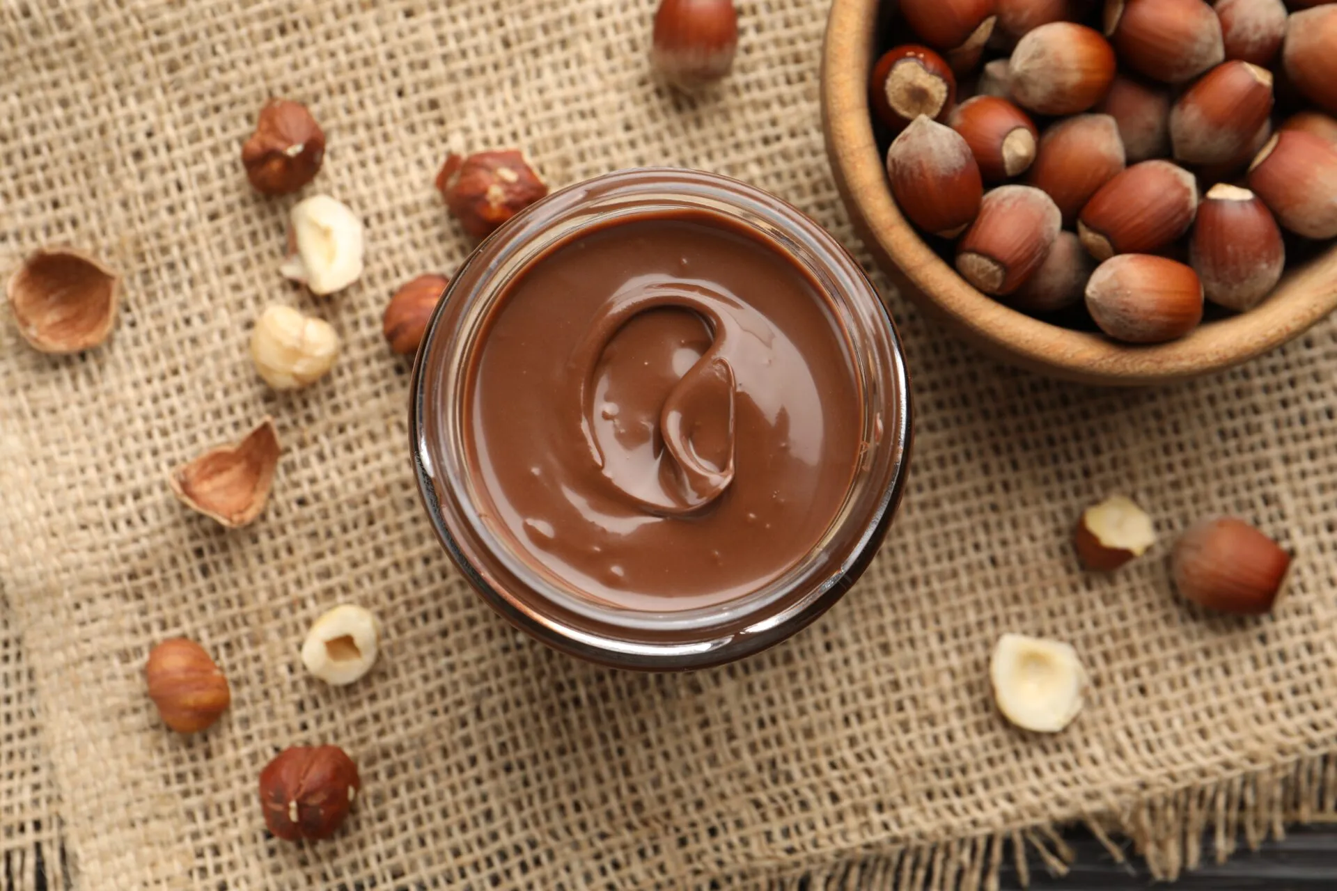 Barattolo di vetro con crema spalmabile alle nocciole vista dall'alto, circondato da nocciole intere e sgusciate su un telo di juta