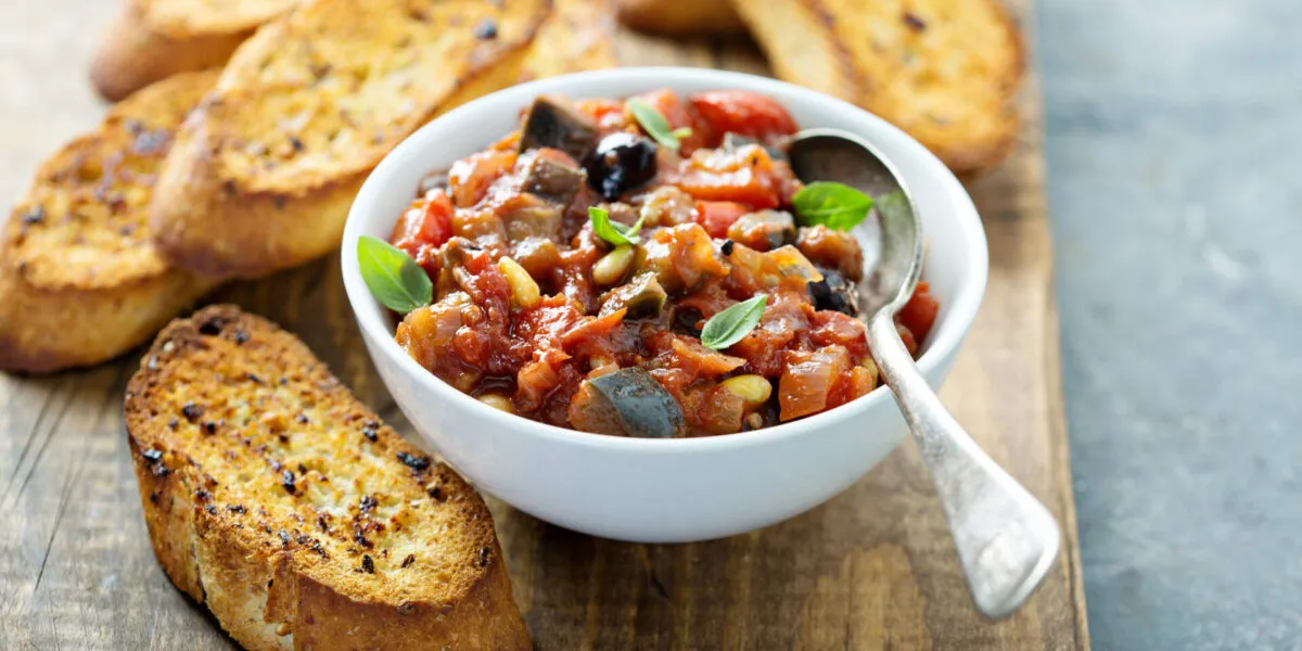 Ciotola bianca di caponata siciliana con melanzane, pomodoro, olive e pinoli, guarnita con basilico, accanto a fette di pane tostato