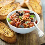 Ciotola bianca di caponata siciliana con melanzane, pomodoro, olive e pinoli, guarnita con basilico, accanto a fette di pane tostato