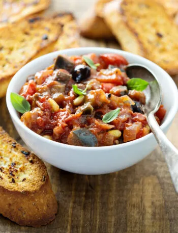 Ciotola bianca di caponata siciliana con melanzane, pomodoro, olive e pinoli, guarnita con basilico, accanto a fette di pane tostato