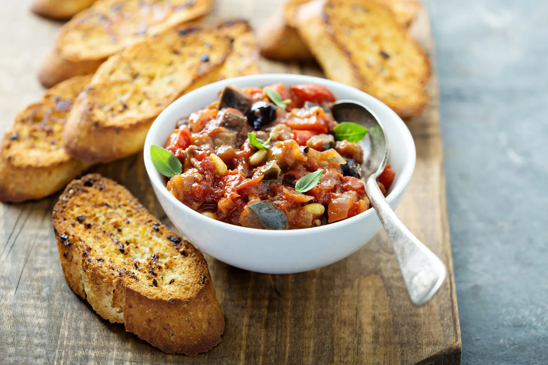 Ciotola bianca di caponata siciliana con melanzane, pomodoro, olive e pinoli, guarnita con basilico, accanto a fette di pane tostato
