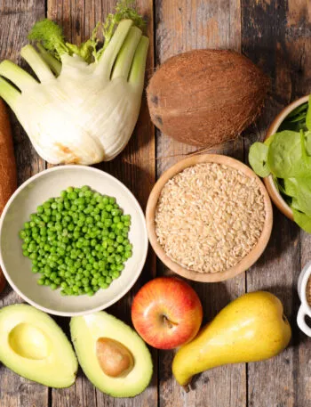 Vista dall’alto di alimenti ricchi di fibre su un tavolo di legno: pane integrale, finocchio, piselli, riso integrale, spinaci, avena, mele, pera e avocado