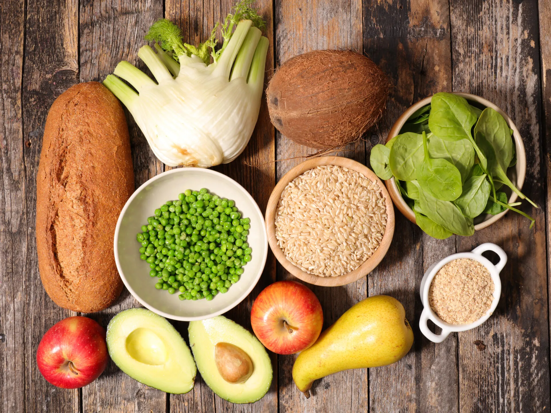 Vista dall’alto di alimenti ricchi di fibre su un tavolo di legno: pane integrale, finocchio, piselli, riso integrale, spinaci, avena, mele, pera e avocado