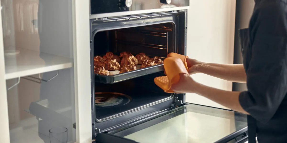 Donna che estrae una teglia di dolcetti dorati da un forno a incasso aperto, con guanto arancione e manopole di funzione e temperatura visibili