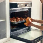 Donna che estrae una teglia di dolcetti dorati da un forno a incasso aperto, con guanto arancione e manopole di funzione e temperatura visibili