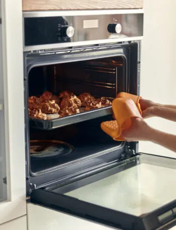 Donna che estrae una teglia di dolcetti dorati da un forno a incasso aperto, con guanto arancione e manopole di funzione e temperatura visibili