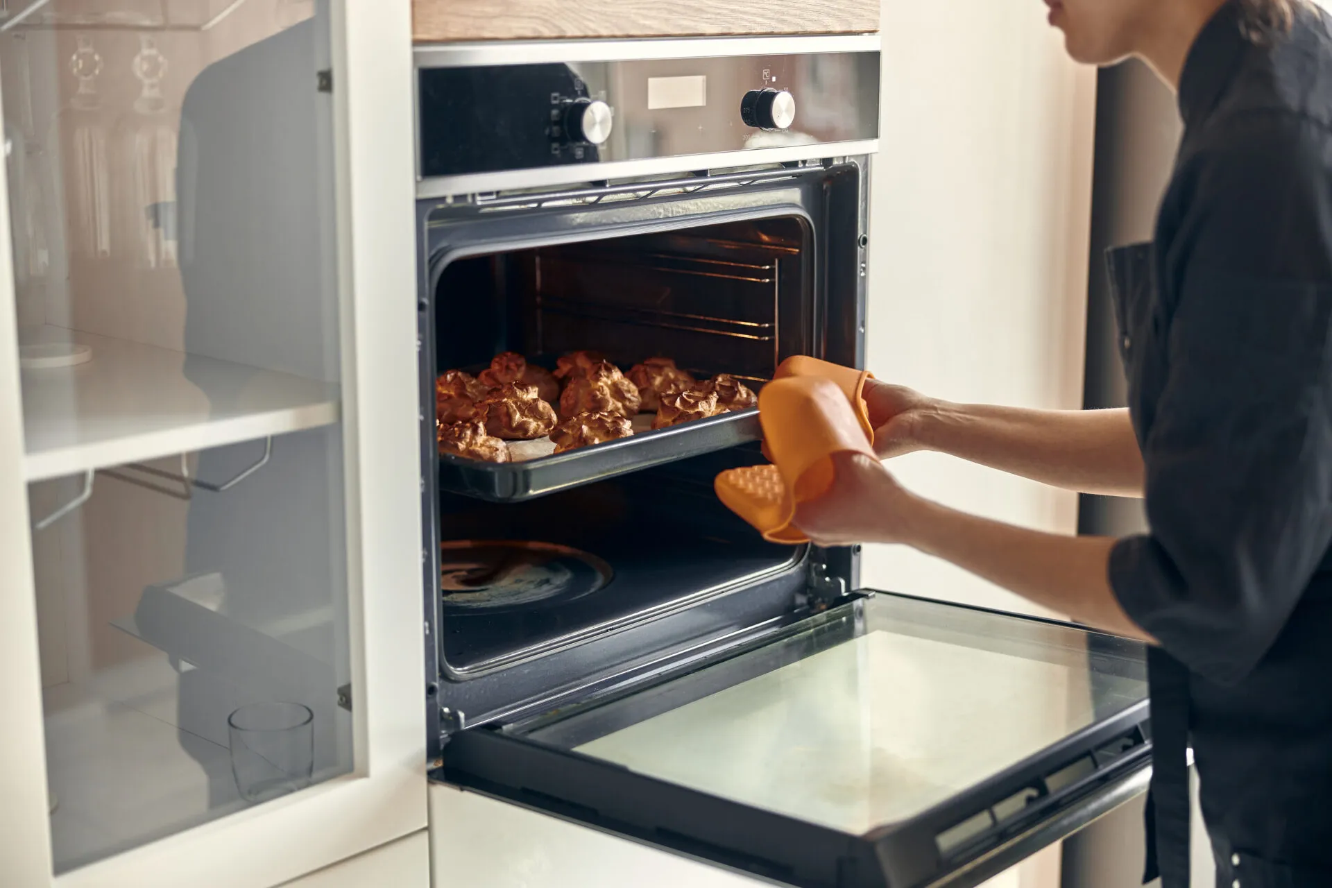 Donna che estrae una teglia di dolcetti dorati da un forno a incasso aperto, con guanto arancione e manopole di funzione e temperatura visibili