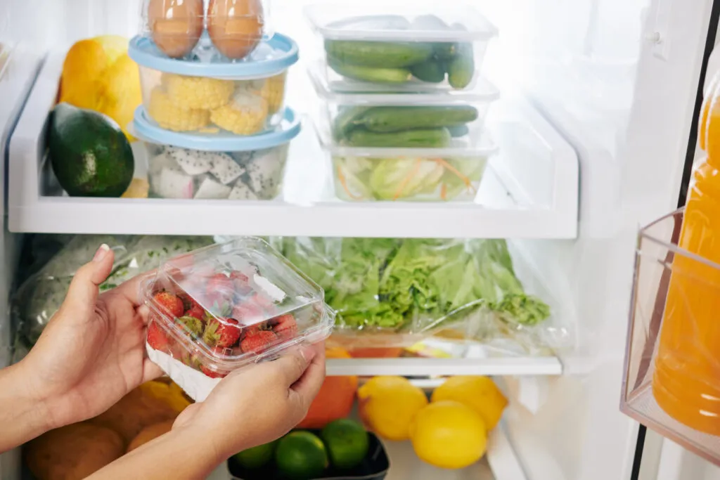 Frigorifero aperto e ordinato con contenitori trasparenti di verdure e frutta, e una mano che prende una vaschetta di fragole