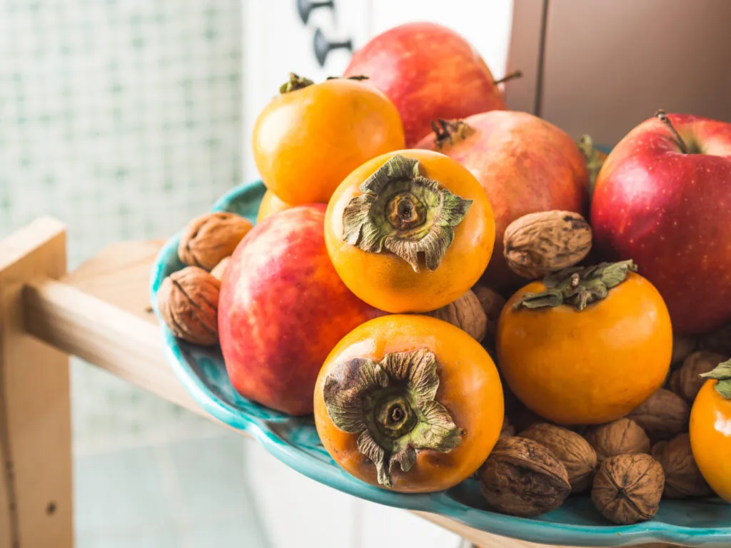 Fruttiera di ceramica turchese con cachi arancioni, mele rosse, una melagrana e noci con il guscio, su una mensola di legno chiaro