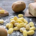 Gnocchi di patate crudi rigati su un piano di legno infarinato, con patate intere e un rigagnocchi di legno accanto